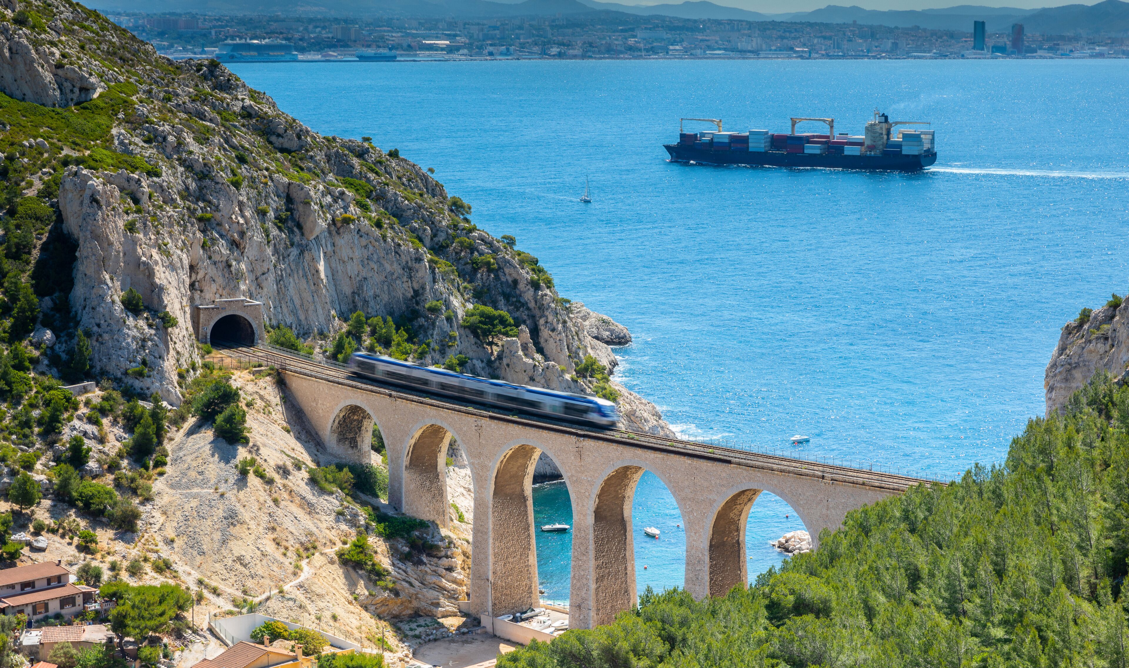 A coastal bridge with a train passing over it, a cargo ship in the water, and a mountainous landscape.