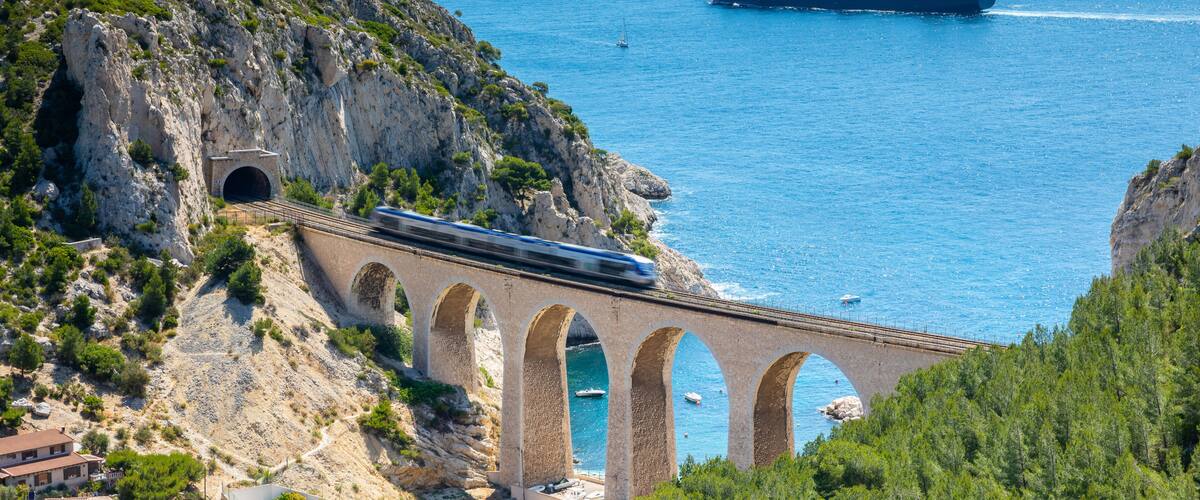 A coastal bridge with a train passing over it, a cargo ship in the water, and a mountainous landscape.