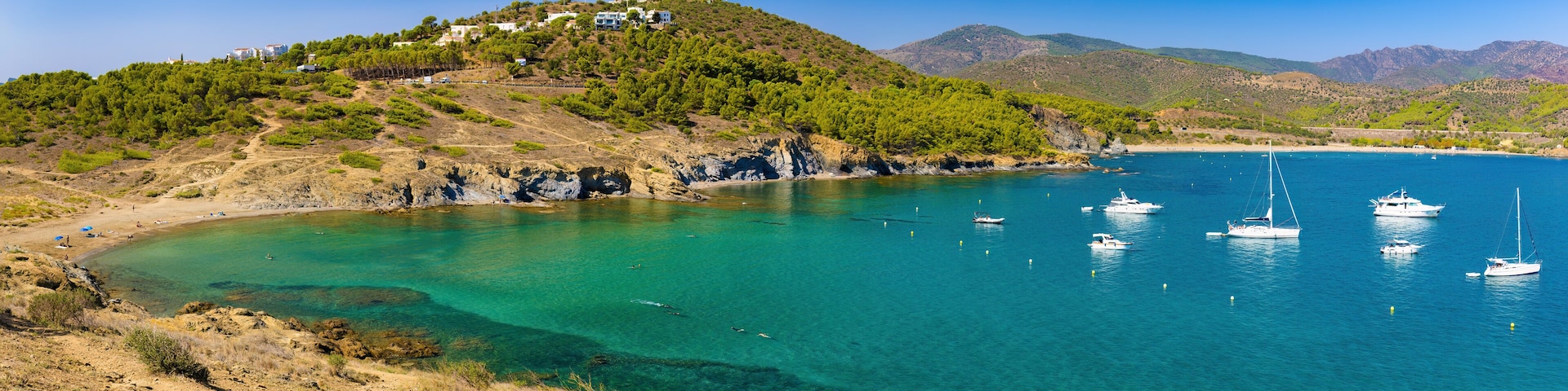 A coastal landscape with a hill covered in greenery, a sandy beach, and several boats anchored in the clear blue waters.