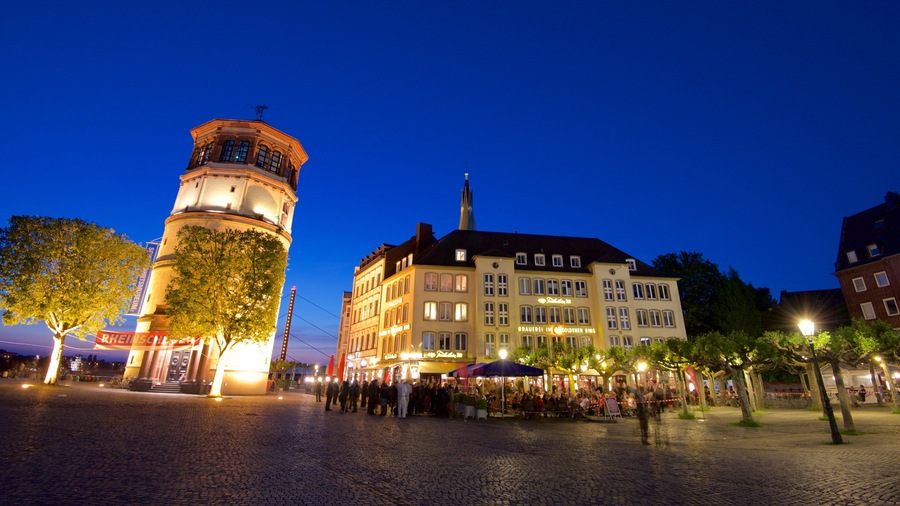 A historic square with cobblestone pavement, a prominent tower, and a large building with illuminated signage.