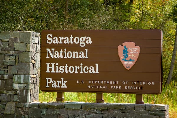 Skilt for Saratoga National Historical Park, med en stenvæg og træer i baggrunden.