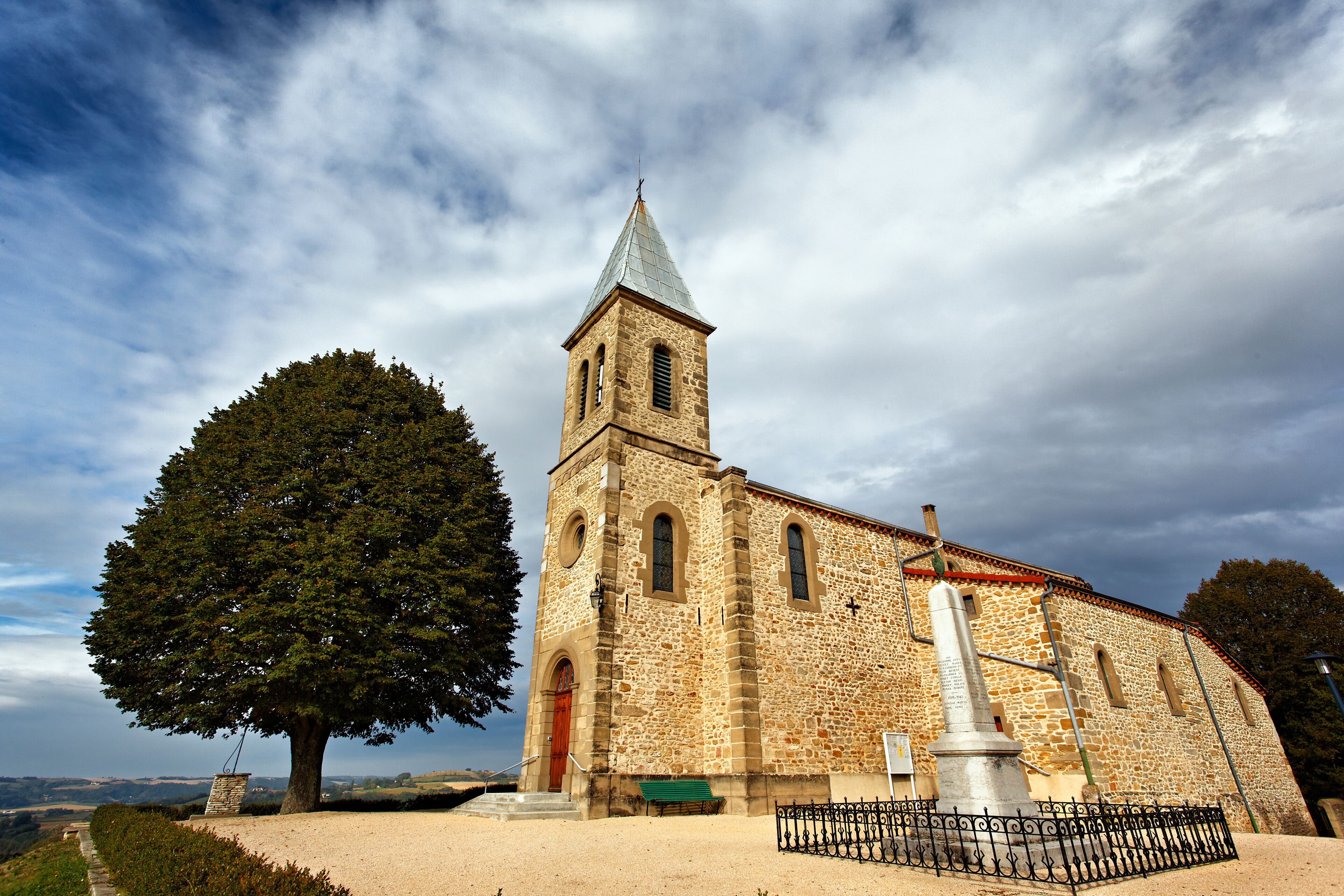 A stone church with a tall tower, a large tree in front, and a metal fence.