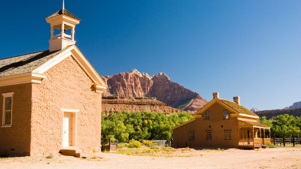 A two-story brick building with a bell tower, a smaller adjacent house, and a mountainous backdrop.
