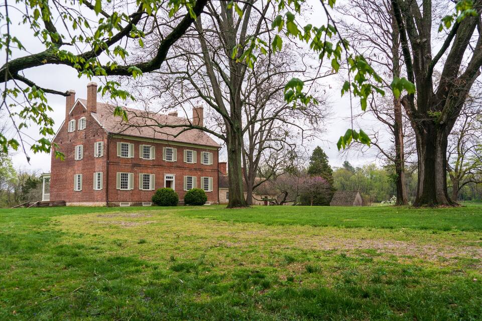 A large brick house with green shutters, surrounded by a grassy field and large trees.