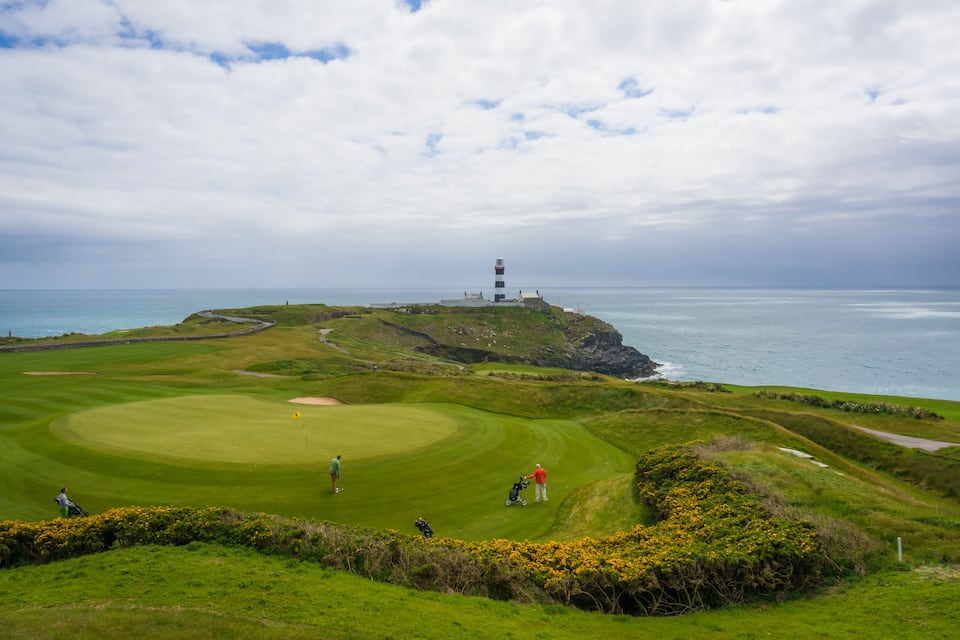 Un parcours de golf situé sur la côte, avec un phare visible au loin, entouré de collines verdoyantes et de l’océan.