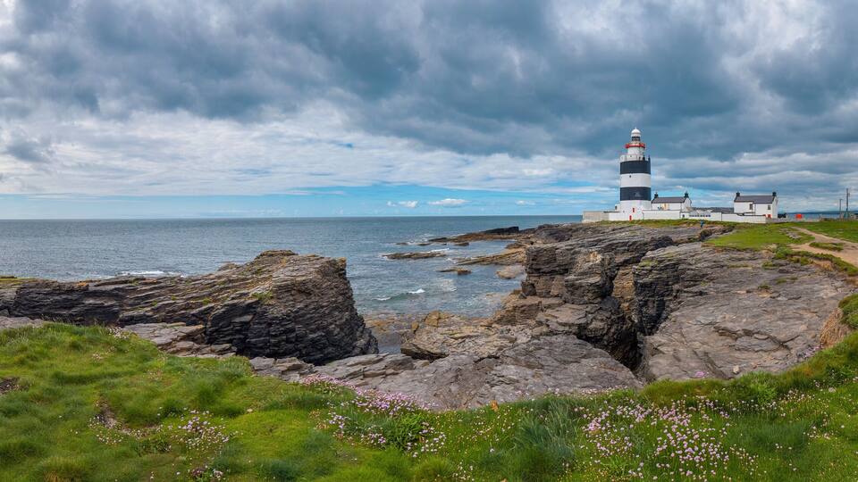 A lighthouse on rocky cliffs by the sea, surrounded by green grass and wildflowers.