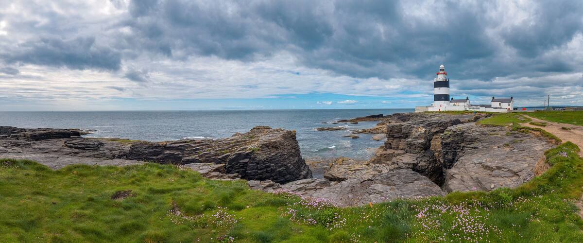 A lighthouse on rocky cliffs by the sea, surrounded by green grass and wildflowers.