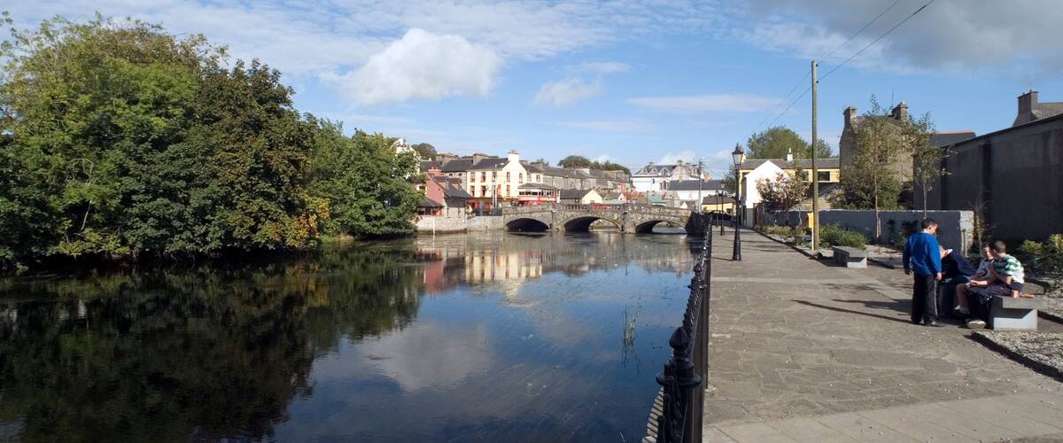 A calm river with a bridge, lush greenery, and buildings along the riverbank.