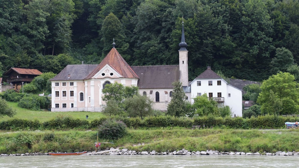 A church with a tall steeple and a bell tower, surrounded by lush greenery and a river.