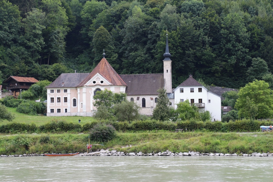A church with a tall steeple and a bell tower, surrounded by lush greenery and a river.