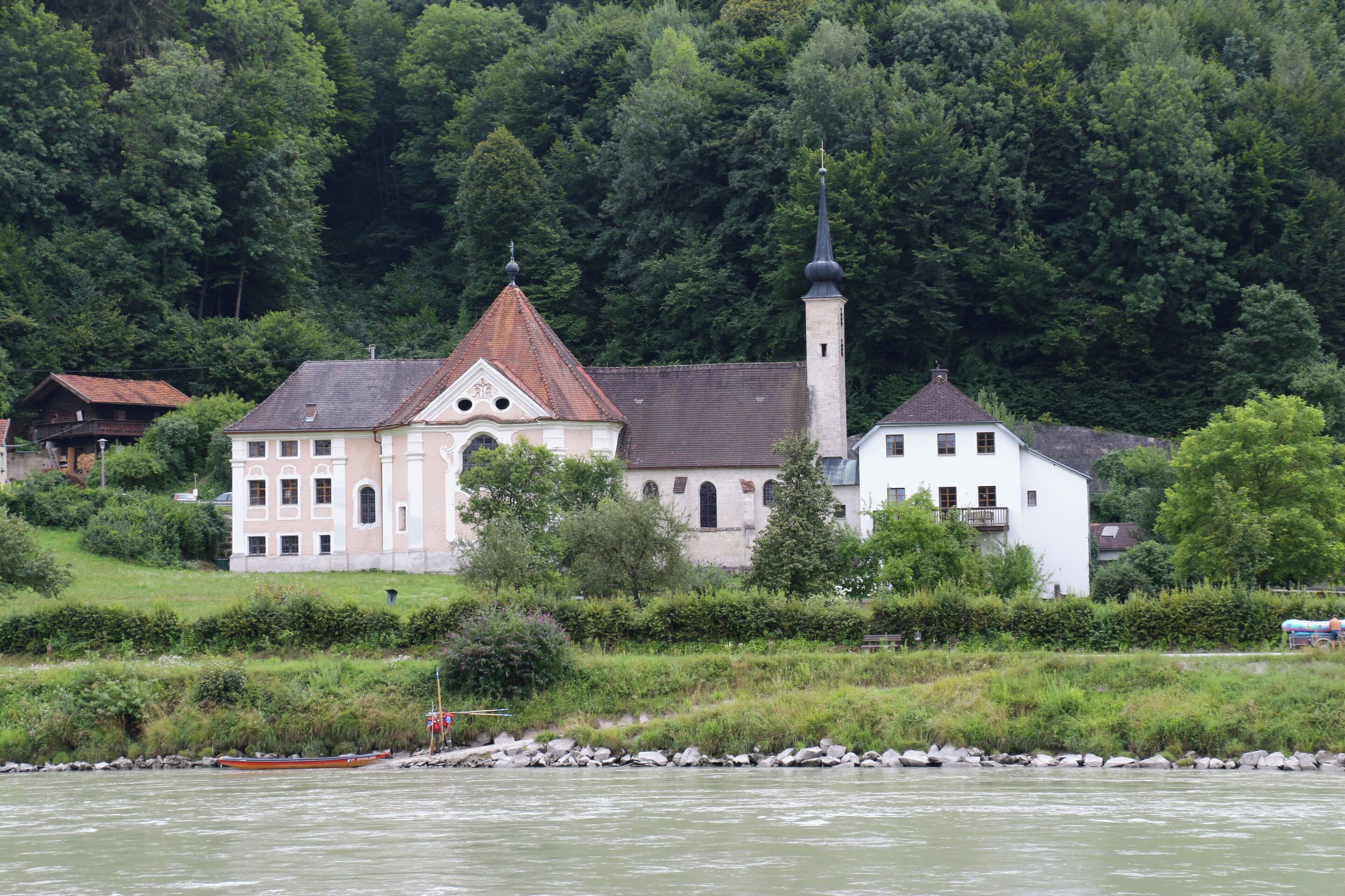 A church with a tall steeple and a bell tower, surrounded by lush greenery and a river.