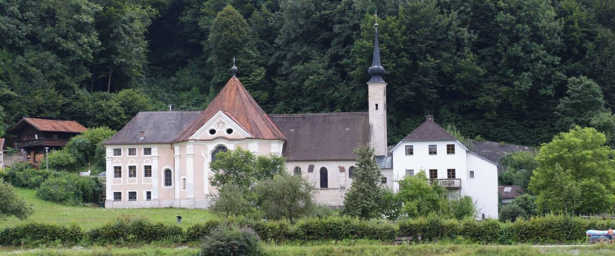 A church with a tall steeple and a bell tower, surrounded by lush greenery and a river.