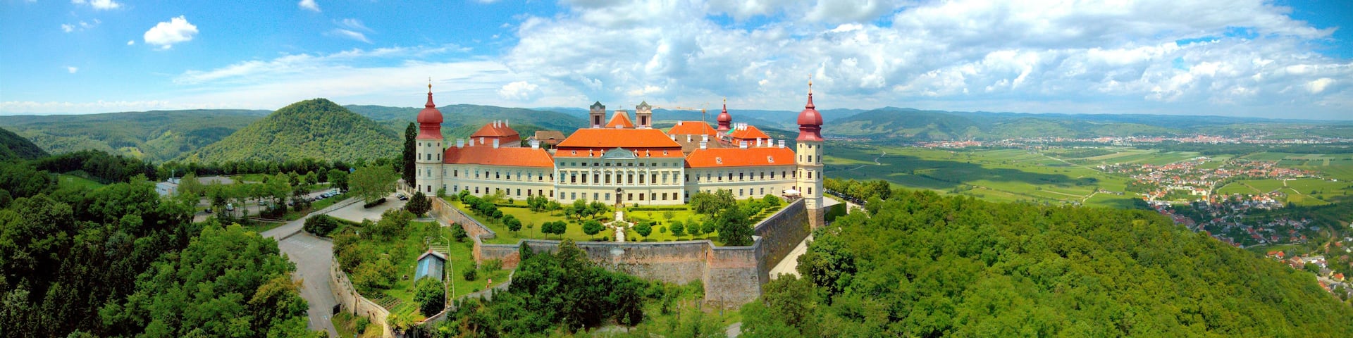 A large, historic building with red domes, surrounded by lush greenery and a mountainous landscape.