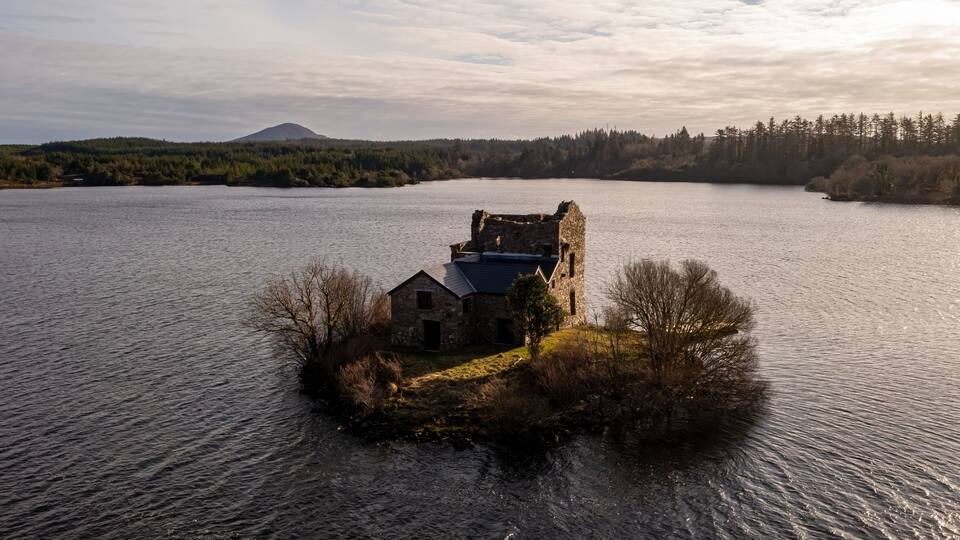 A stone ruin on an island in a lake, surrounded by forest and a mountain in the distance.