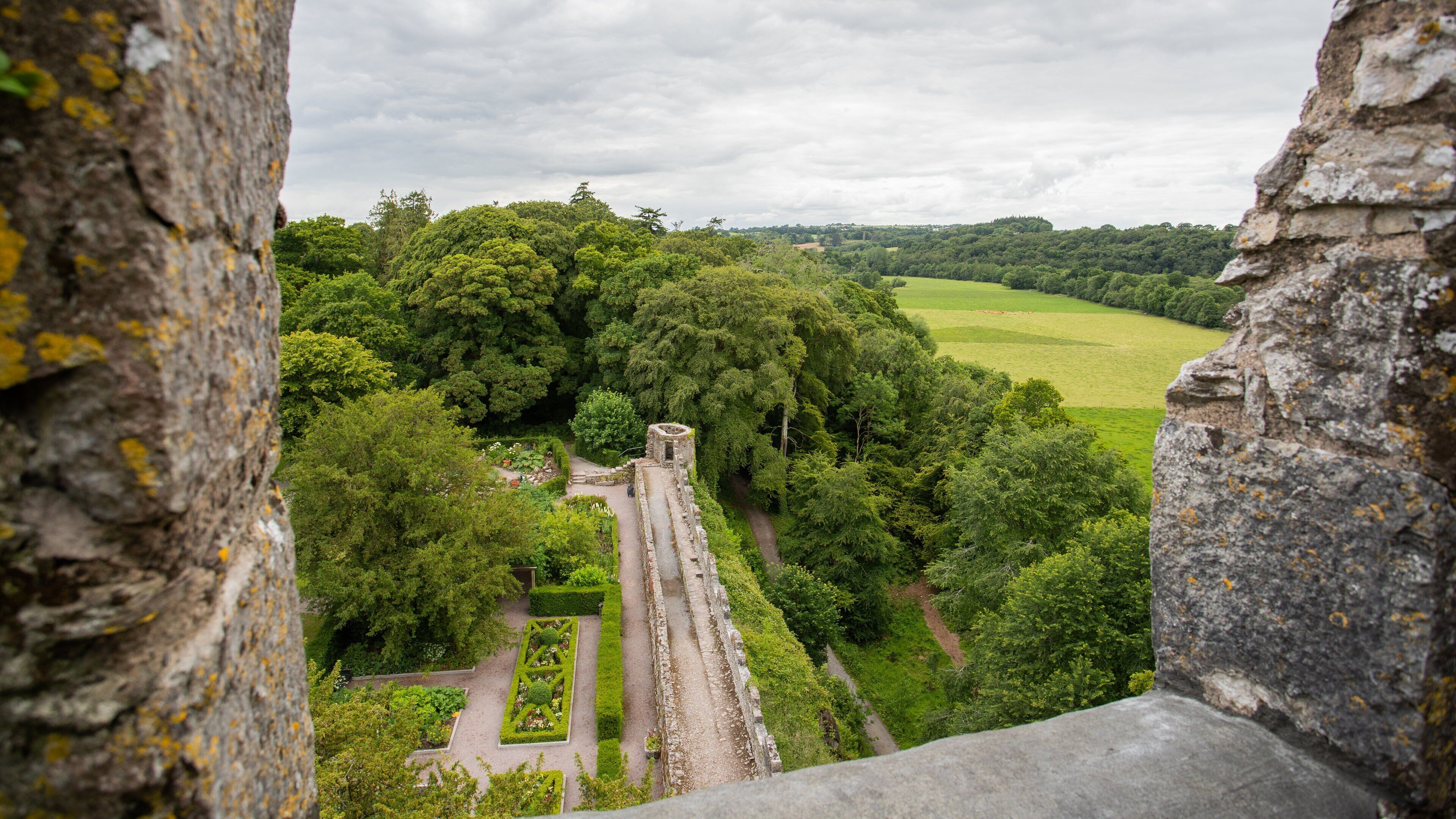 A historical stone structure surrounded by lush greenery and a distant view of a green field.