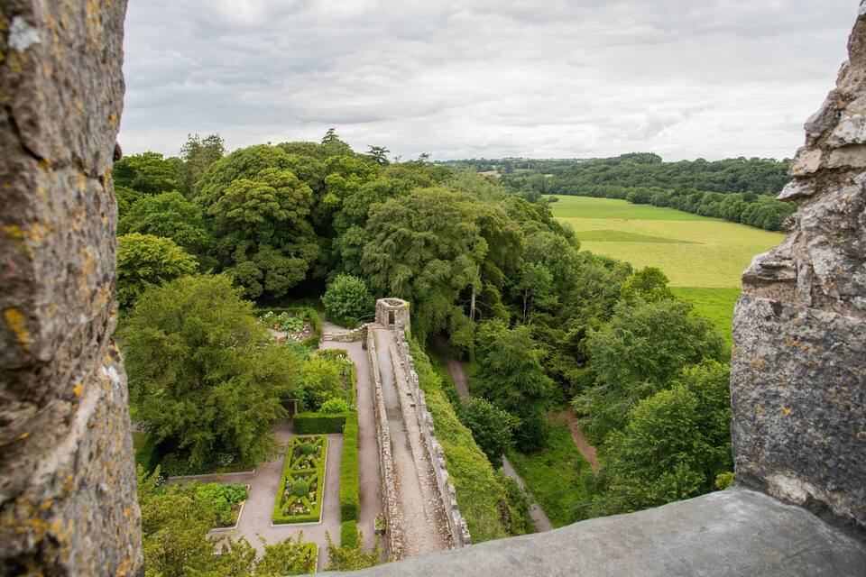 A historical stone structure surrounded by lush greenery and a distant view of a green field.