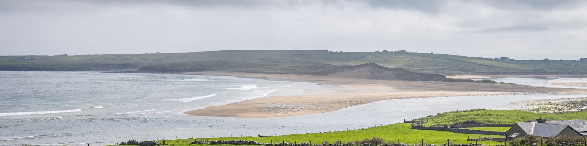 A coastal landscape with a grassy foreground, a sandy beach, and a stone wall.