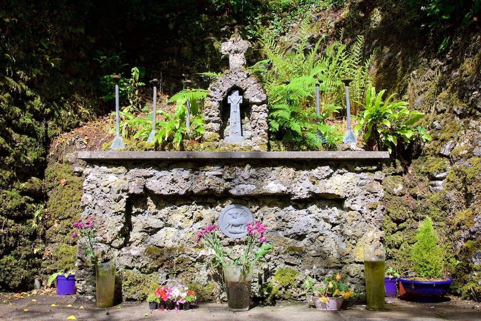 Un santuario de piedra con una cruz, rodeado de helechos y flores, frente a un muro de roca cubierto de musgo.