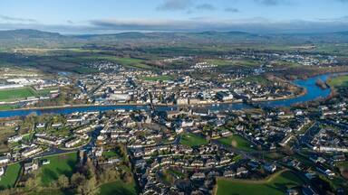 An aerial view of a town with a river running through it, surrounded by green fields and hills.
