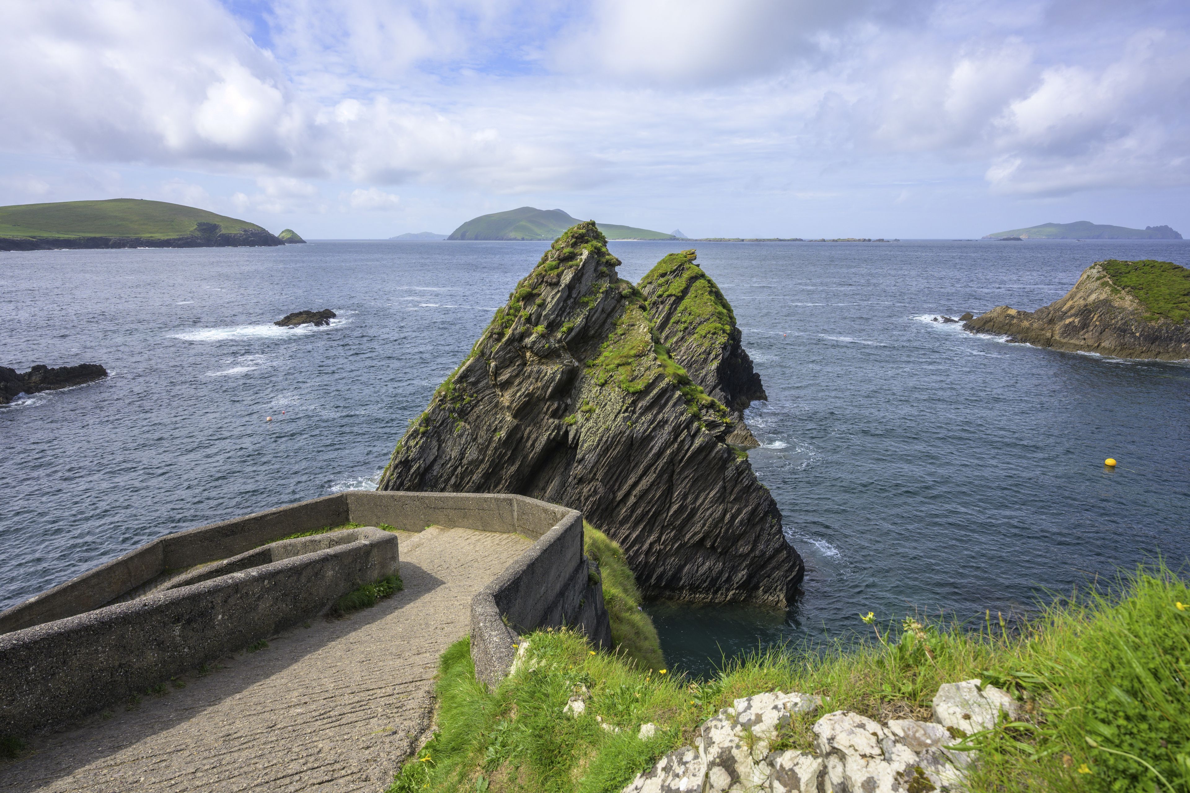 A coastal landscape with a prominent rock formation, a sea wall, and distant islands under a partly cloudy sky.