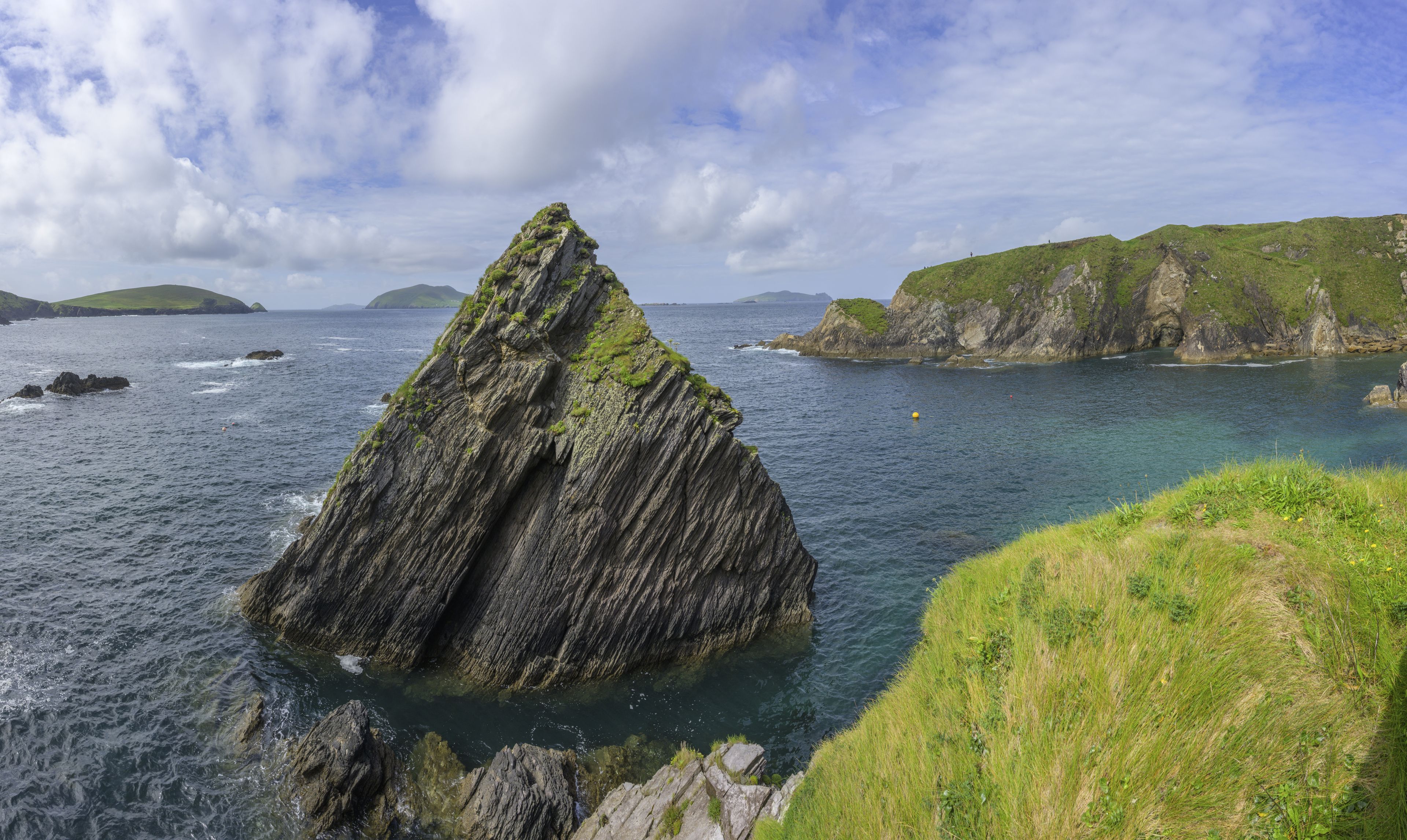 A rugged coastline with a prominent rock formation surrounded by calm sea and grassy hills.