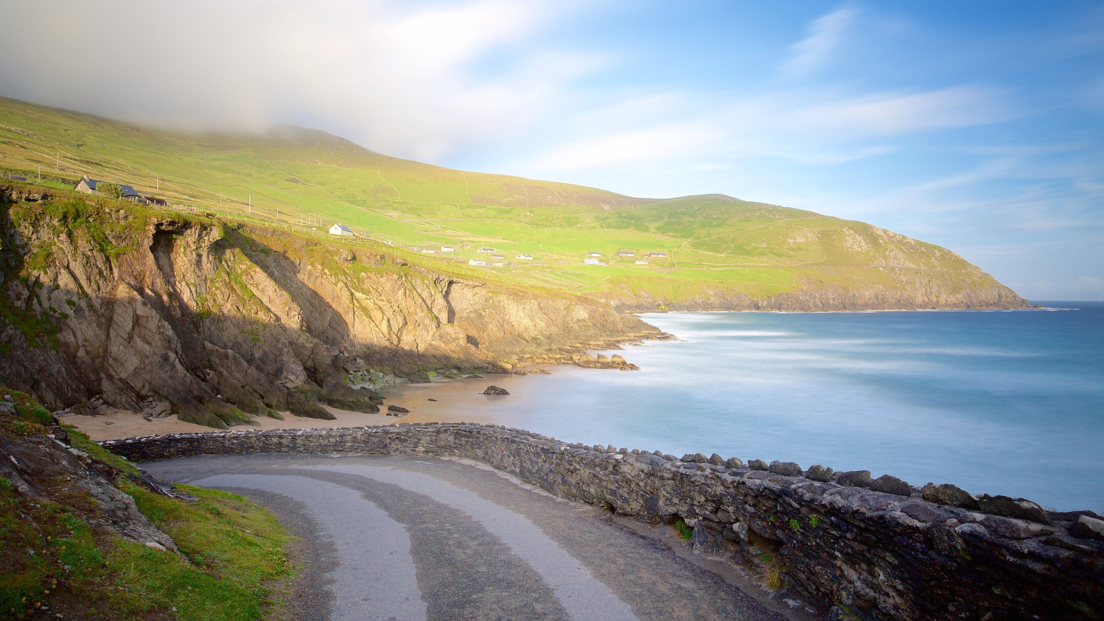 A coastal road winding through a rocky landscape with a view of the sea and distant houses on a green hillside.