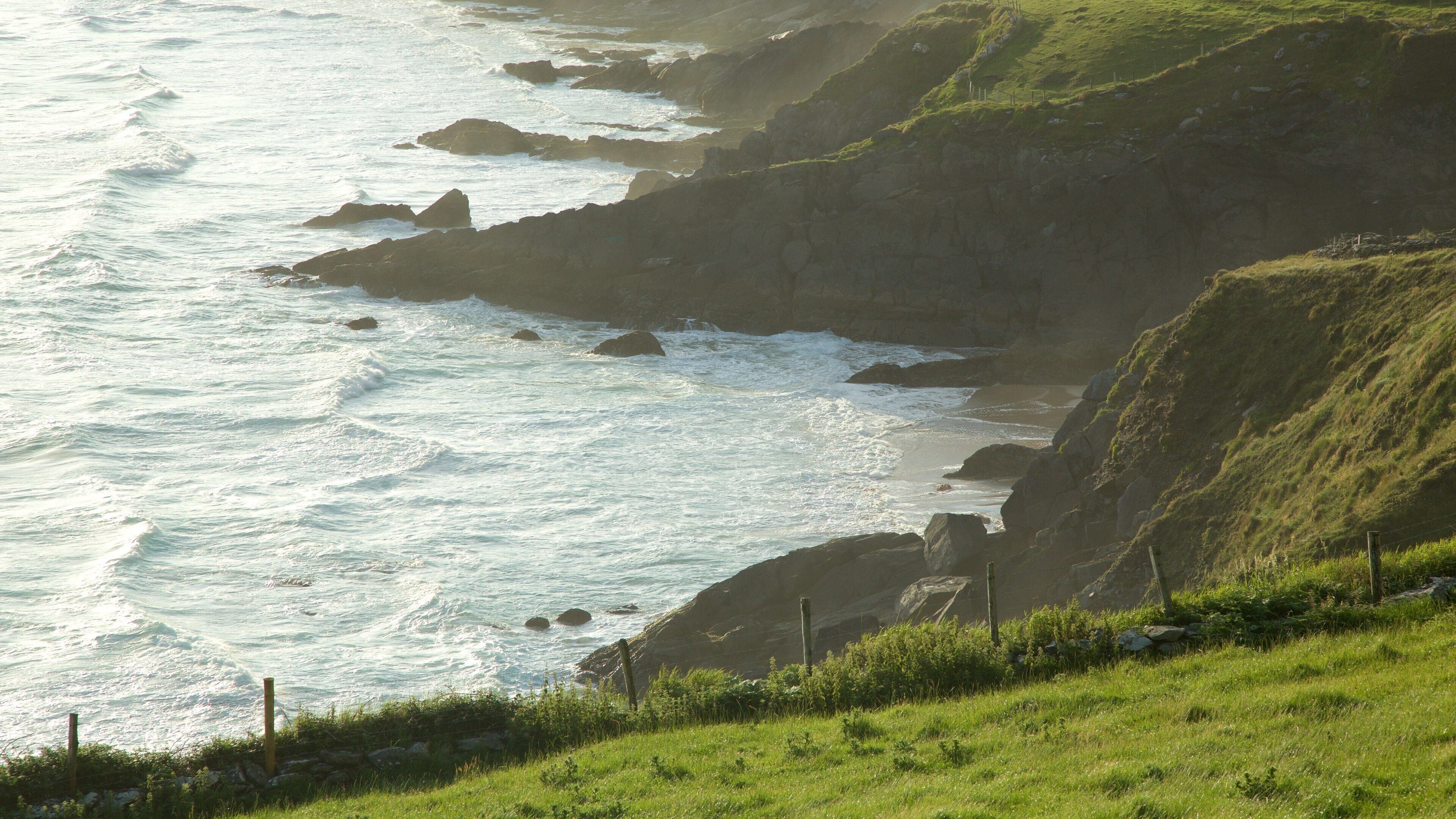 A coastal landscape with waves crashing against rocky cliffs and a grassy hillside.