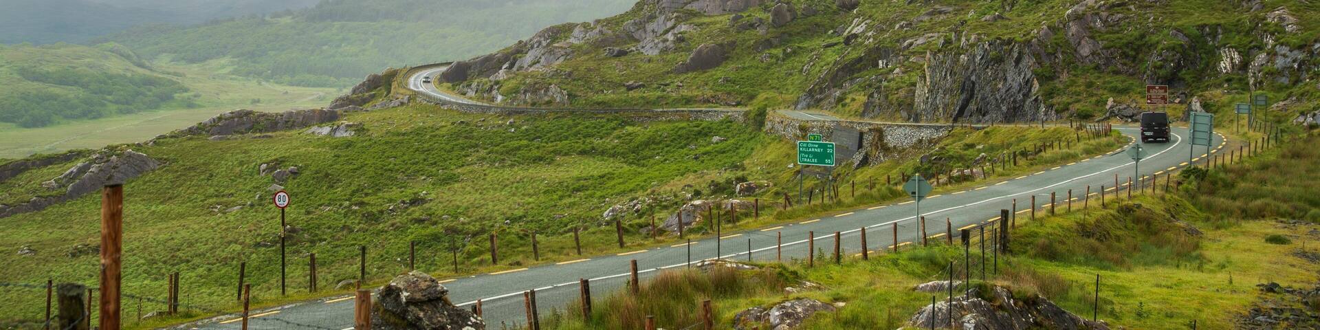 A winding mountain road with a car, surrounded by lush green hills and rocky outcrops.