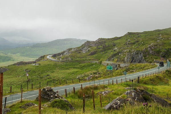 Une route de montagne sinueuse, avec une voiture, bordée de collines verdoyantes et de formations rocheuses.