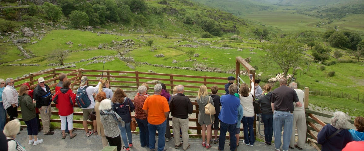 A group of people standing on a wooden fence overlooking a green valley with mountains in the background.