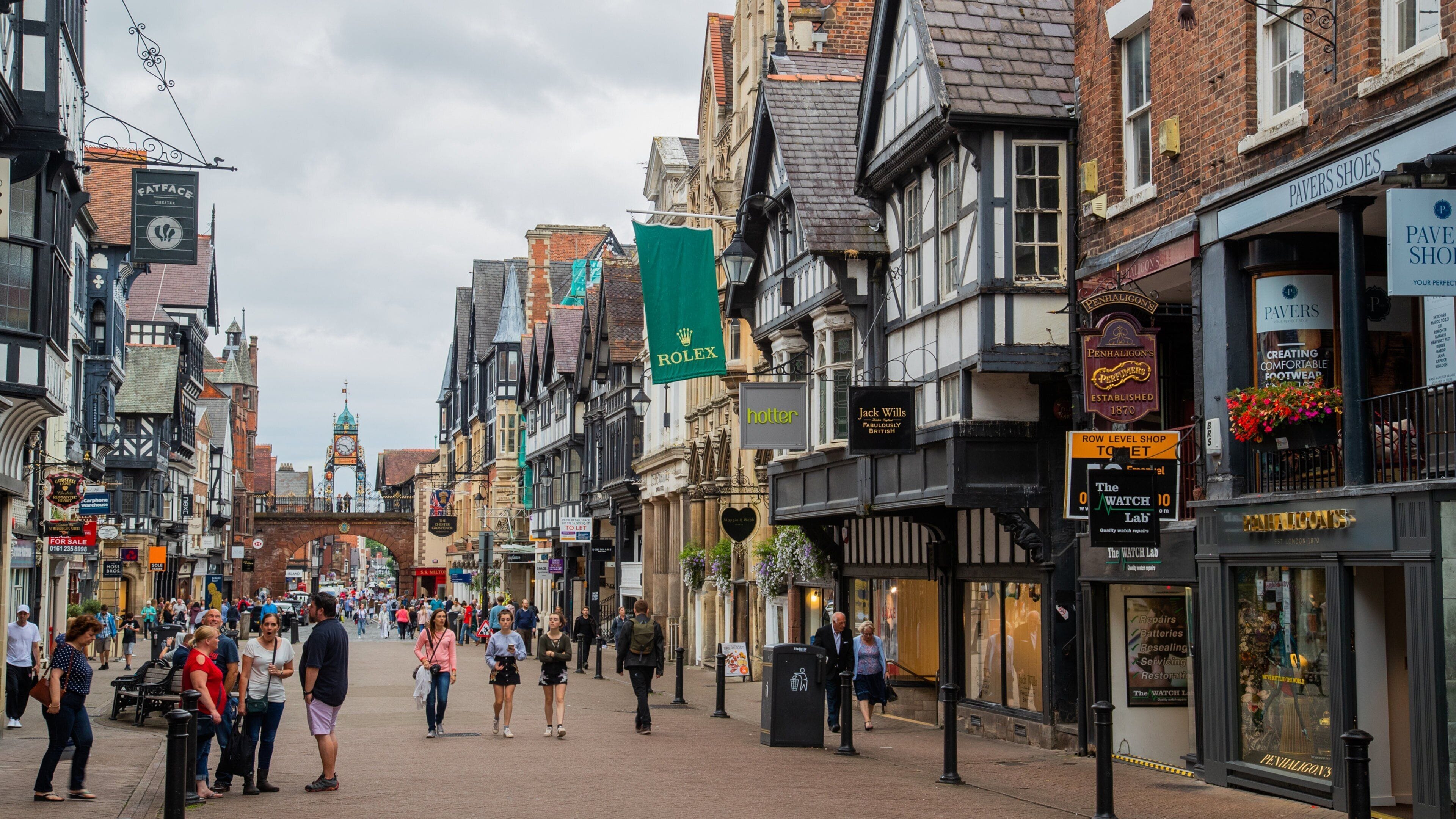 Una calle concurrida en un pueblo histórico con edificios de estilo Tudor, tiendas y una imponente torre del reloj.
