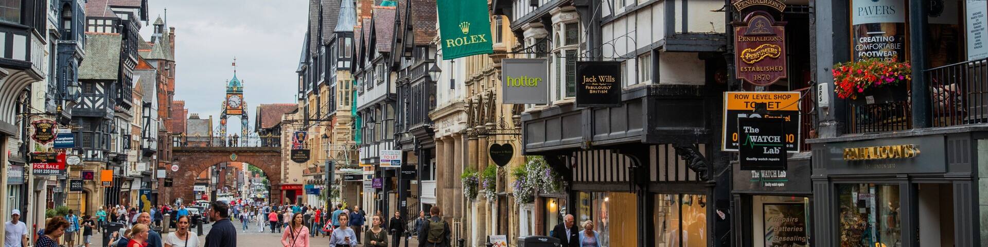 A busy street in a historic town with Tudor-style buildings, shops, and a prominent clock tower.