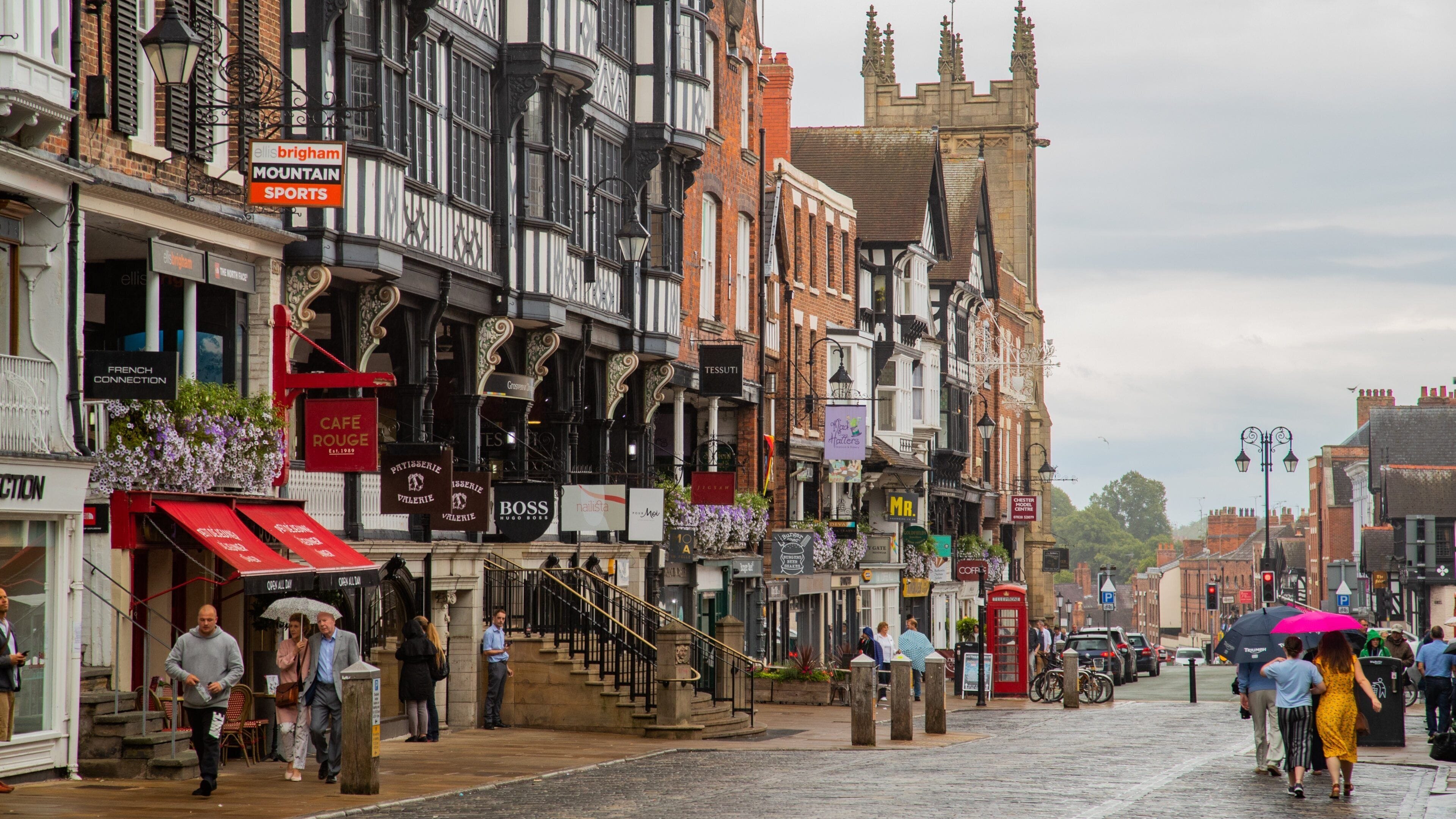 Una calle histórica con edificios de estilo Tudor, comercios y una imponente torre del reloj.