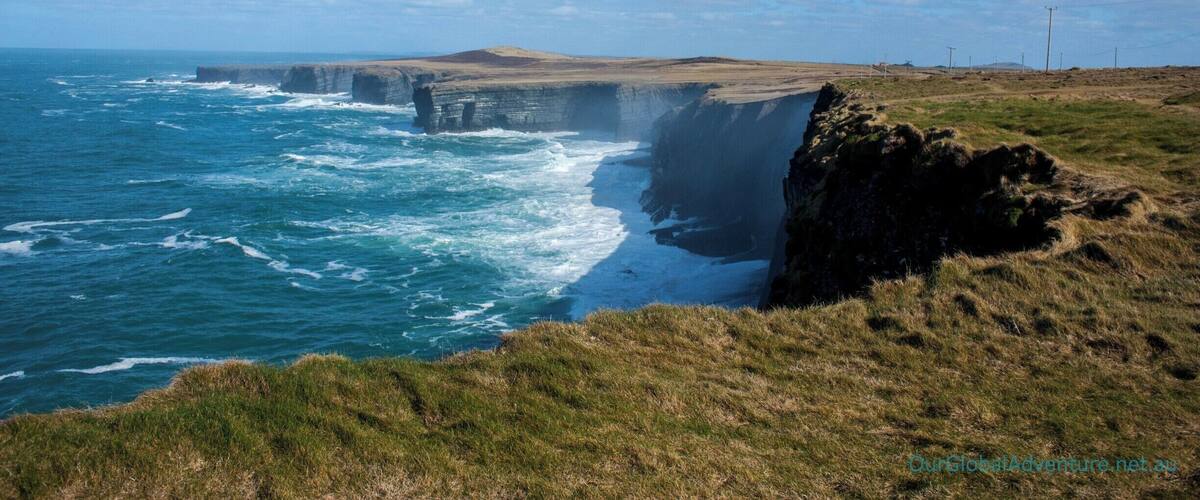 A coastal cliff with waves crashing against it, a grassy landscape, and a clear sky.