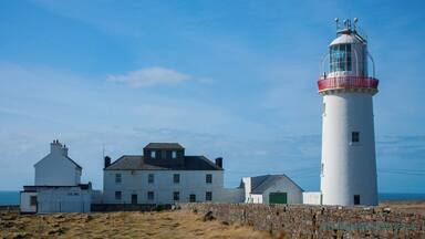 A lighthouse with a white and red top, a white building with a green door, and a stone wall in front.