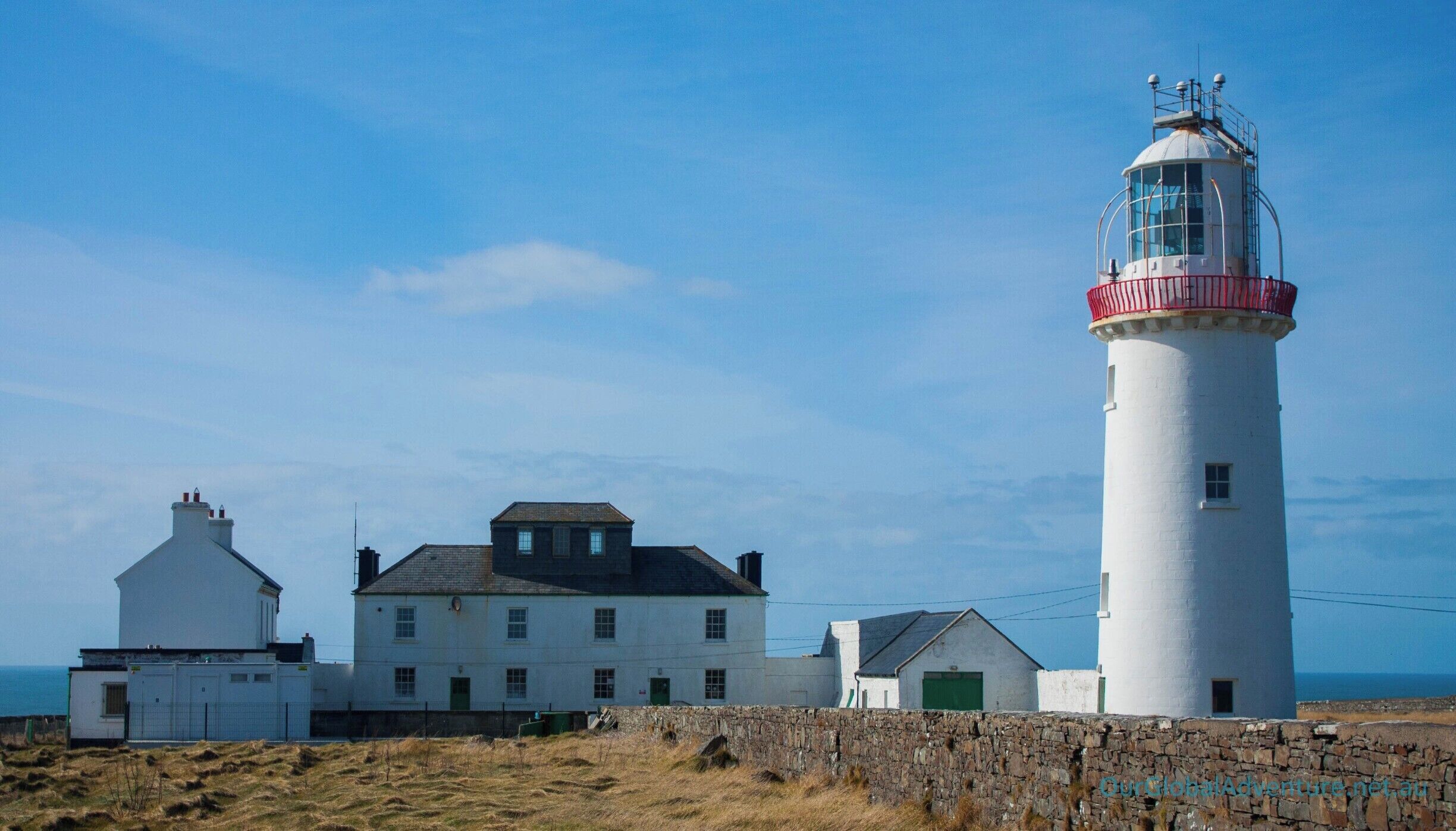A lighthouse with a white and red top, a white building with a green door, and a stone wall in front.