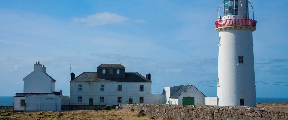 A lighthouse with a white and red top, a white building with a green door, and a stone wall in front.