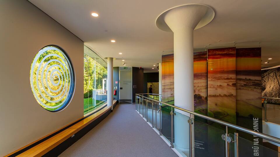 A modern building interior with a glass railing, a large circular artwork, and a mural depicting a sunset landscape.