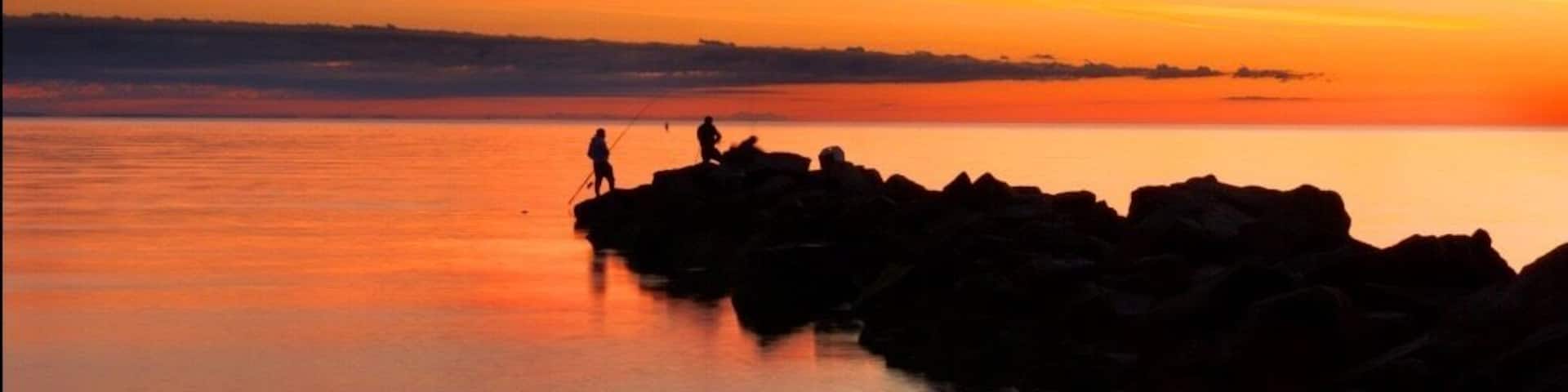 A sunset over a calm body of water with rocky outcrops and silhouettes of people fishing.