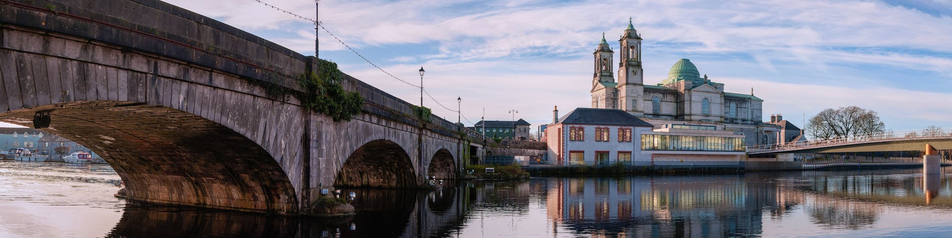 A stone bridge over a calm river, reflecting a historic building with two towers and a green dome.