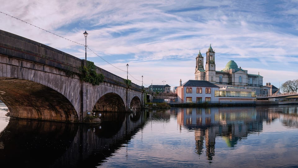 A stone bridge over a calm river, reflecting a historic building with two towers and a green dome.