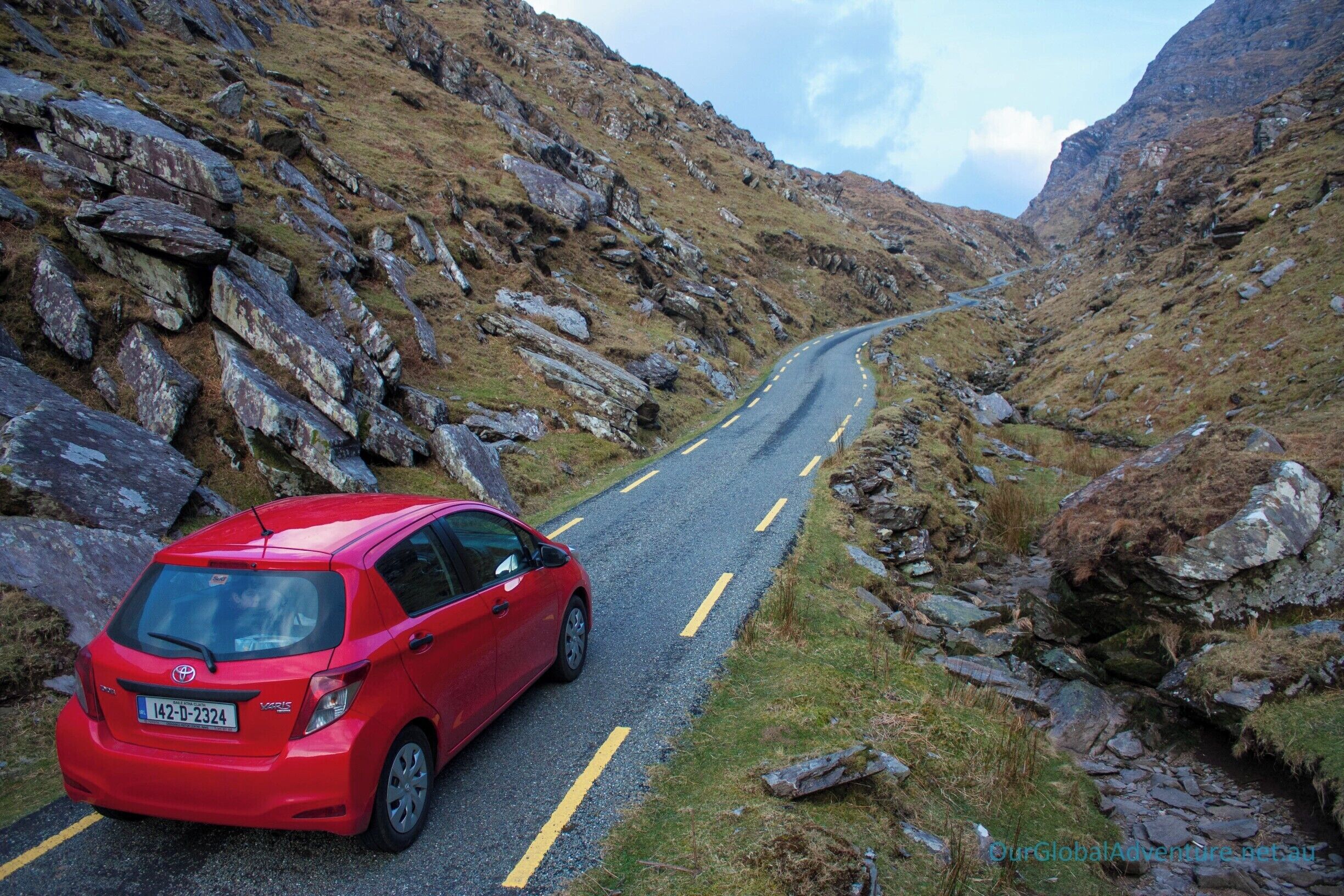 Un coche rojo en una estrecha carretera de montaña rodeado de terreno rocoso.