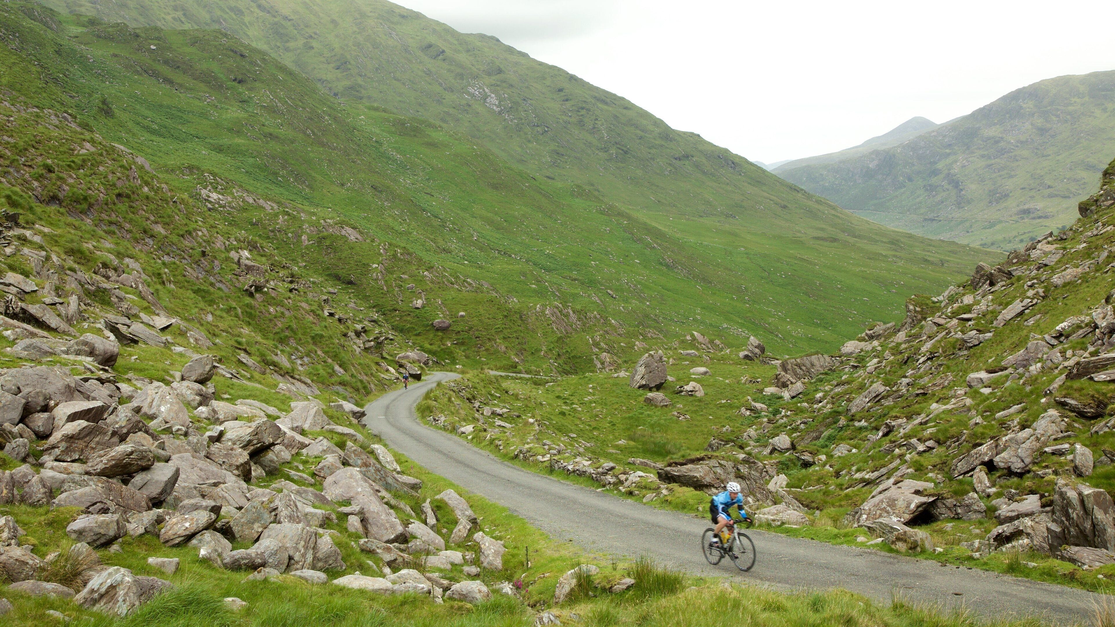 A cyclist on a winding mountain road surrounded by rocky terrain and lush green hills.