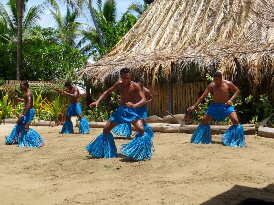 A group of people performing a dance in a sandy area with a thatched-roof hut in the background.