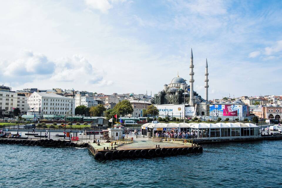 A waterfront cityscape with a prominent mosque featuring two minarets and a dome, a large white building with a blue sign, and a covered structure with yellow railings.