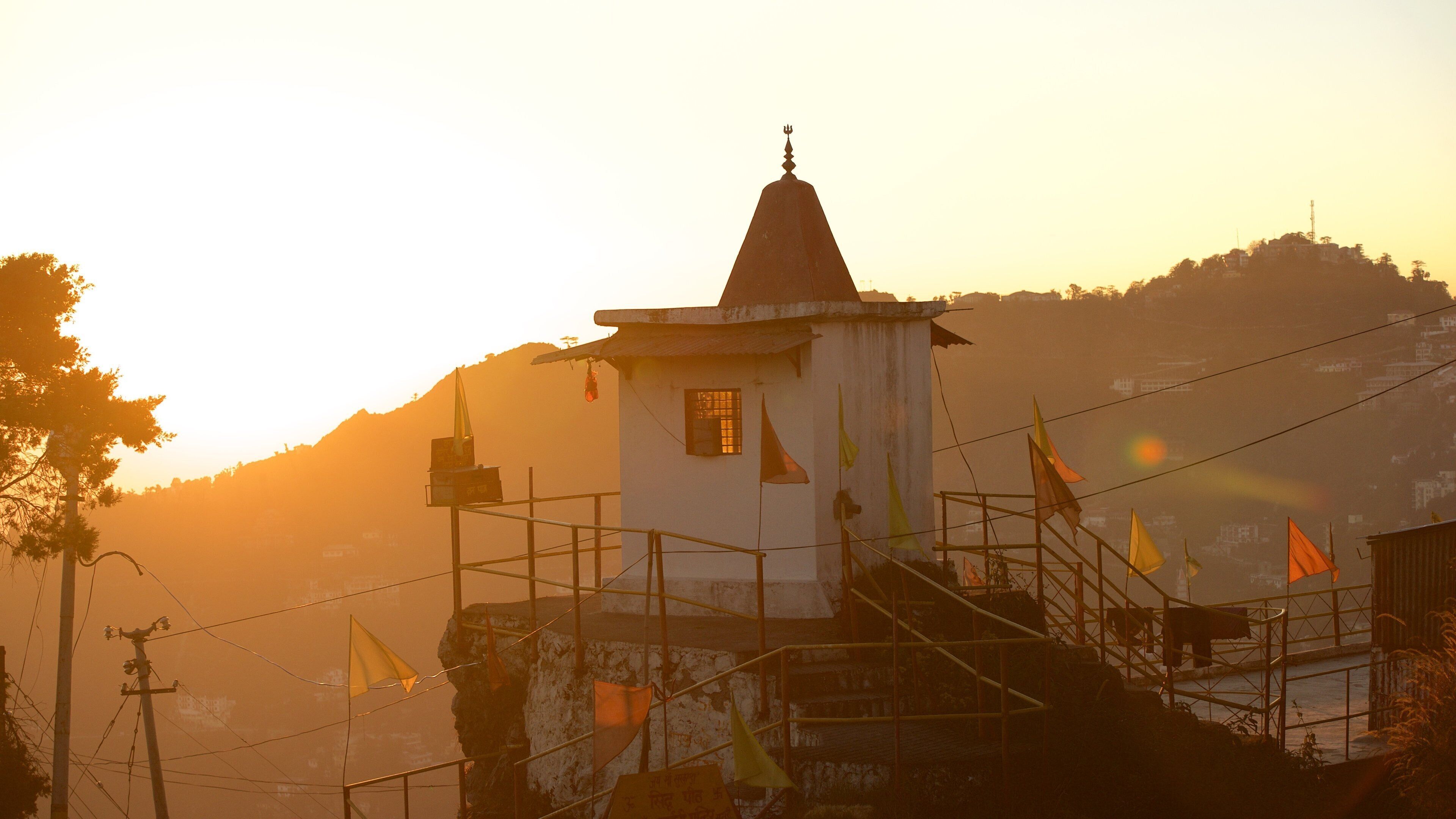 A small temple with a spire, surrounded by flags and a mountainous backdrop during sunset.