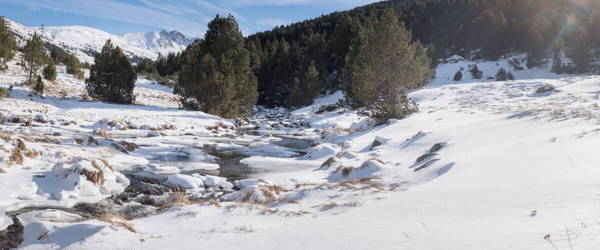 A snow-covered landscape with a river, trees, and mountains in the background.