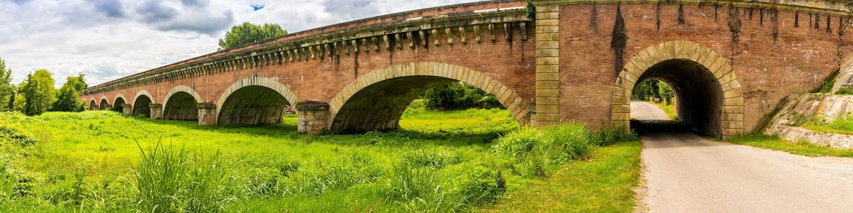 Eine Ziegelbrücke mit mehreren Bögen über einer Straße, umgeben von üppigem Grün und Bäumen.