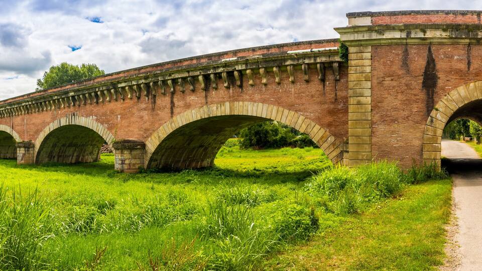 A brick viaduct with multiple arches over a road, surrounded by lush green grass and trees.