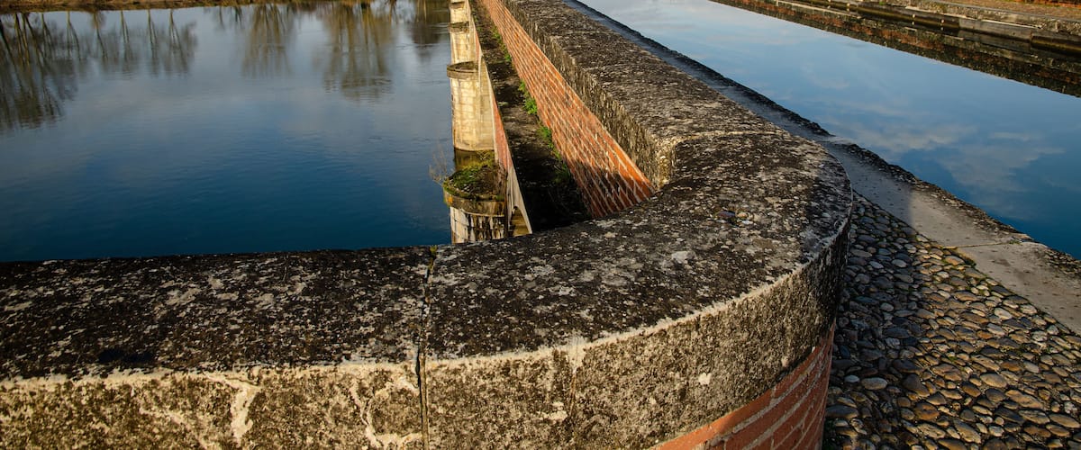 A canal with a brick wall and calm water reflecting the sky.